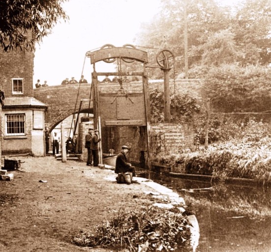 One of the Guillotine Lock Gates at Lifford Lane Bridge on the Stratford-upon-Avon Canal. The building to the left is the toll collector’s office. The lock gates were located about 350 yards from the end of the canal where it joins the Worcester Canal. This first section of the canal was completed to Lapworth by 1802. Since the nationalisation of the canals in 1948, water loss from one canal to another is less of an issue, so the gates are no longer in use and are both left open. The last recorded use of the gates was 1959. This photo shows how fascinated the English are by canals – there are spectators in abundance on the bridge and the canal side even in these early days. Or are they supposed to be working?